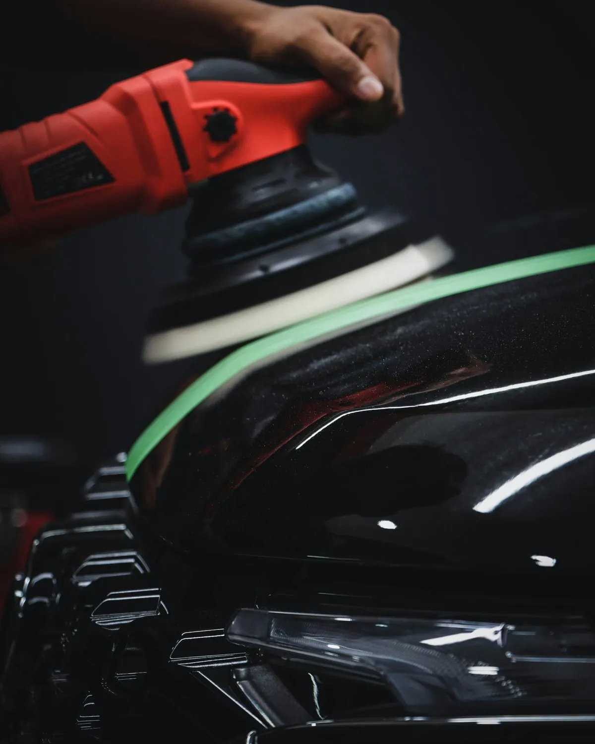 Detailer using a red orbital polisher on a black car bonnet with green masking tape